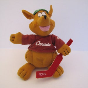 May include: A brown plush kangaroo wearing a maroon shirt with "Canada" embroidered on the front and a green hat. The kangaroo is holding a red hockey stick with "ROOTS" printed on it.