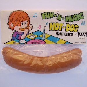 May include: A brown plastic harmonica shaped like a hot dog. The packaging features a cartoon illustration of a child eating a hot dog and the text "Fun-N-Music Hot-Dog Harmonica WY No. 831 Made in Hong Kong".