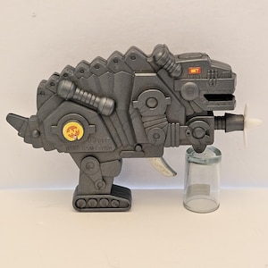 May include: A gray plastic toy water gun shaped like a dinosaur. The toy has a propeller on the front and a small glass jar attached. The text "WING HANG TOYS" is visible on the side.