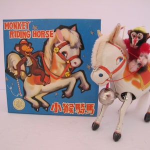 Peut inclure: Jouet en tôle vintage représentant un cheval blanc avec une selle rouge et orange, monté par un singe en tenue rouge. Le jouet a une cloche argentée et est à côté d'une boîte bleue avec l'inscription "MONKEY RIDING HORSE".