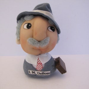 May include: A vintage doll of a man in a grey suit, hat, and tie. The doll has a light skin tone, a grey moustache, and the text "L.M. DeBoss" on the front. A small brown briefcase is attached.