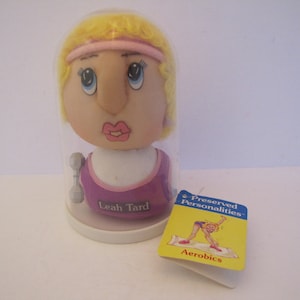 May include: A plastic dome encases a plush doll head with blonde hair, wearing a pink headband and a purple and pink exercise outfit. The doll's face has blue eyes, a pink nose, and a pink mouth. The base of the dome has the text "Leah Tard" and a yellow tag with the text "Preserved Personalities - Aerobics" and a cartoon image of a woman doing aerobics.