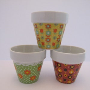 May include: Three ceramic flower pots with different floral patterns. The top pot has a yellow background with red and green flowers. The middle pot has a green background with orange and white polka dots. The bottom pot has a red background with black and yellow flowers.