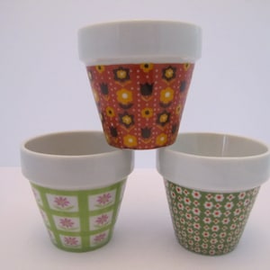 May include: Three small white ceramic flower pots with different floral patterns. The pots are decorated with red, orange, yellow, green, white, and pink flowers.