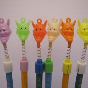 May include: Seven colorful plastic pencil toppers shaped like a Japanese demon mask with a spring attached to the bottom. The pencil toppers are in the colors purple, green, orange, white, orange, yellow, and green.