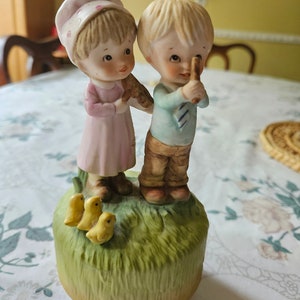 May include: A ceramic figurine featuring a boy and girl standing together on a grassy base, with three small yellow chicks. The girl wears a pink dress and bonnet, while the boy has a blue shirt and brown pants. The figurine is painted in soft pastel colors.