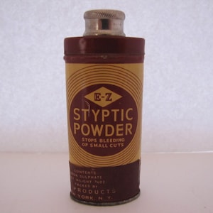 Peut inclure: Un ancien récipient cylindrique de poudre styptique E-Z. Le récipient est rouge foncé avec une étiquette jaune portant le nom du produit et un logo en forme de losange. Le texte indique "Arrête les saignements des petites coupures."