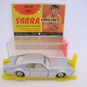 May include: A silver die-cast toy car model of a 1970s Oldsmobile Toronado, labeled "Sabra Super Car" on the box. The car is on a yellow plastic display stand with a clear plastic case.