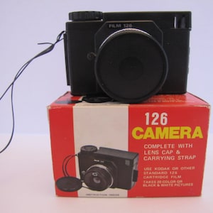 May include: A black 126 camera with a lens cap and carrying strap. The camera is in its original box, which is red with white stripes and has the text "126 CAMERA" in yellow letters. The box also includes instructions for using the camera.