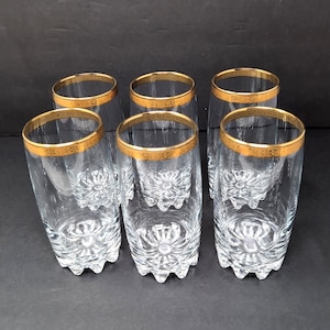 May include: A set of six clear glass tumblers, each featuring a thick gold rim with an intricate etched pattern. The base of each glass has a heavy, starburst-like cut design that radiates outward, providing a textured appearance to the bottom.