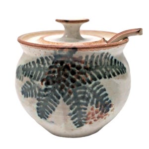 May include: A round ceramic sugar bowl with a speckled off-white glaze, featuring a hand-painted dark green fern leaf pattern. The bowl includes a matching lid with a central knob handle and a small ceramic spoon resting in a side notch.