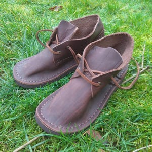 May include: A pair of brown leather ankle boots with a lace-up design. The boots have a rounded toe and are stitched around the sole. The boots are sitting on green grass. The hashtag #handmadeTREK is visible in the image.
