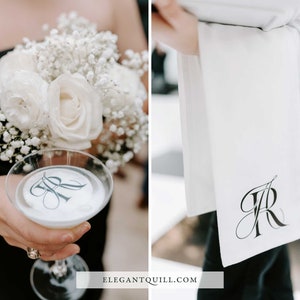 May include: A white cocktail glass with a monogrammed sticker on the bottom. The glass is being held by a person wearing a black dress and holding a bouquet of white roses and baby's breath. A white towel with a monogrammed design is in the background.
