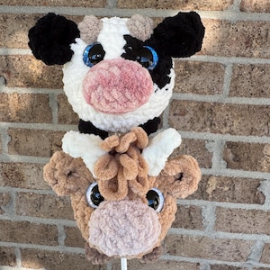Puede incluir: Dos cabezas de vaca de peluche en un palo blanco. Una es blanca y negra con ojos ribeteados en azul y una nariz rosa. La otra es marr&oacute;n con ojos negros y una nariz rosa. Los peluches est&aacute;n contra una pared de ladrillos.