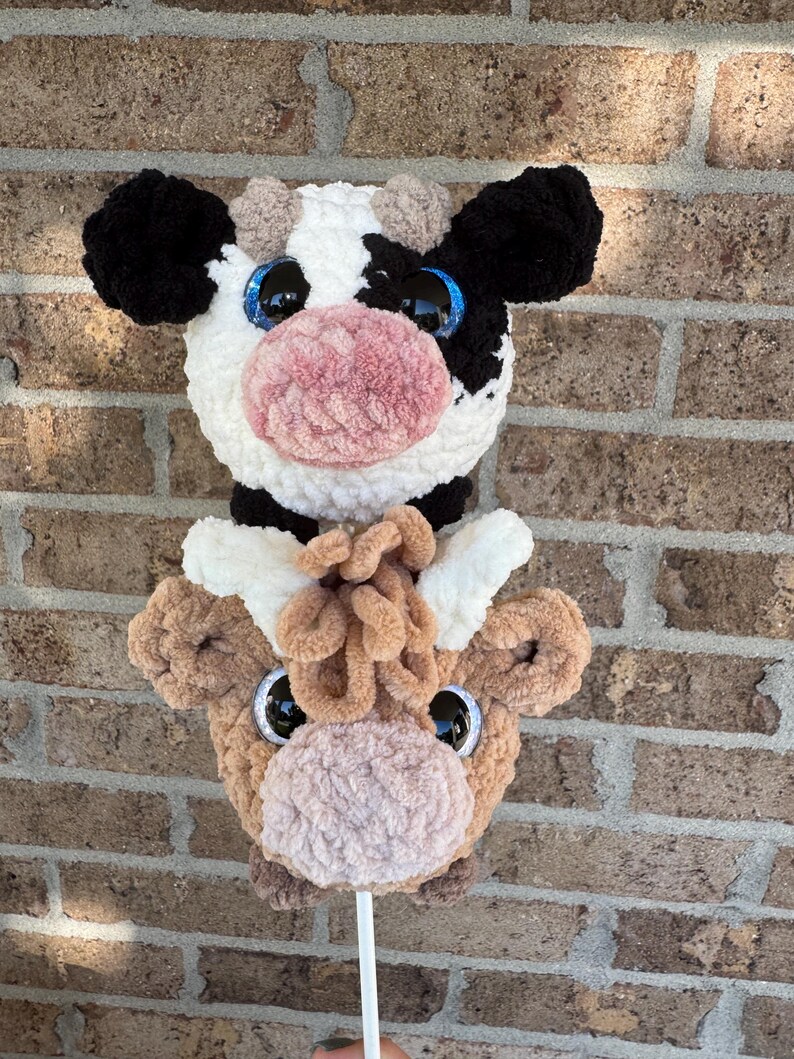 Puede incluir: Dos cabezas de vaca de peluche en un palo blanco. Una es blanca y negra con ojos ribeteados en azul y una nariz rosa. La otra es marr&oacute;n con ojos negros y una nariz rosa. Los peluches est&aacute;n contra una pared de ladrillos.