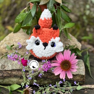 May include: A handmade crochet fox with orange, white, and black details. The fox has large, shiny eyes and is surrounded by flowers and greenery. The Cozy Buckle logo is visible.
