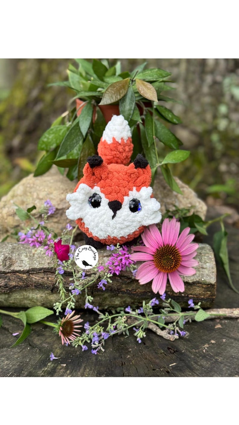 May include: A handmade crochet fox with orange, white, and black details. The fox has large, shiny eyes and is surrounded by flowers and greenery. The Cozy Buckle logo is visible.