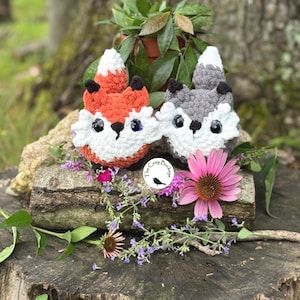 May include: Two crocheted fox plushies, one orange and white, the other gray and white, sit on a weathered log. The foxes have black eyes and noses. The Cozy Bunch logo is visible. The scene is decorated with flowers and greenery.