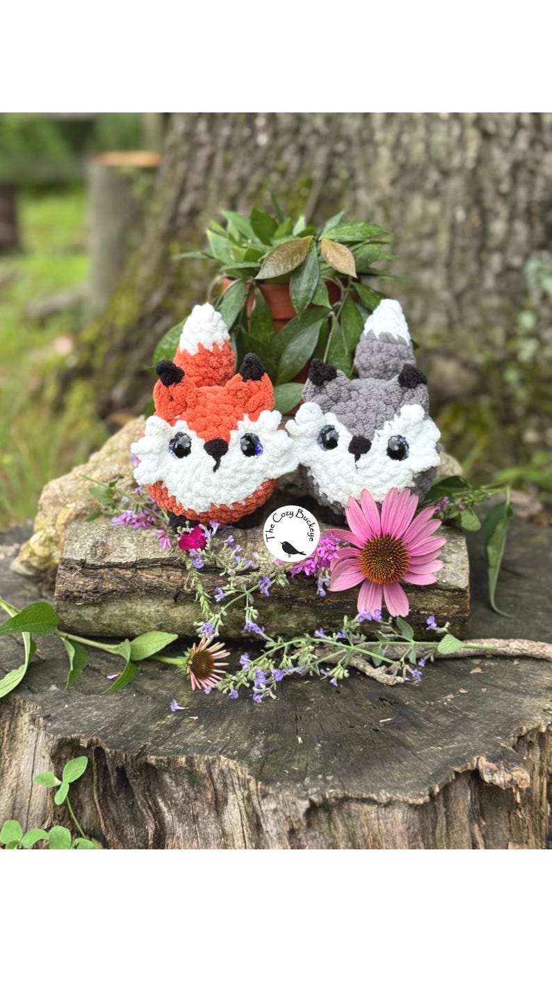 May include: Two crocheted fox plushies, one orange and white, the other gray and white, sit on a weathered log. The foxes have black eyes and noses. The Cozy Bunch logo is visible. The scene is decorated with flowers and greenery.