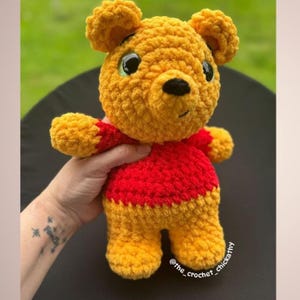 May include: A handmade crochet Winnie the Pooh plush toy. The bear is primarily yellow with a red shirt. It has black eyes and a black nose. The toy is held in a hand. The text "@the_crochet_chickathy" is visible.