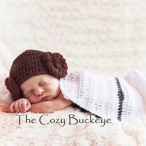 May include: A baby wearing a brown crocheted hat with a white crocheted blanket. The hat has a bun on top and the blanket has a black stripe.