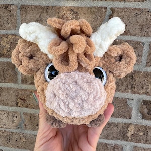 Puede incluir: Cabeza de vaca de peluche hecha a mano con ojos negros grandes y brillantes. La vaca tiene un cuerpo marr&oacute;n claro, una nariz color crema y cuernos color crema. El peluche se sostiene en una mano sobre un fondo de ladrillos.