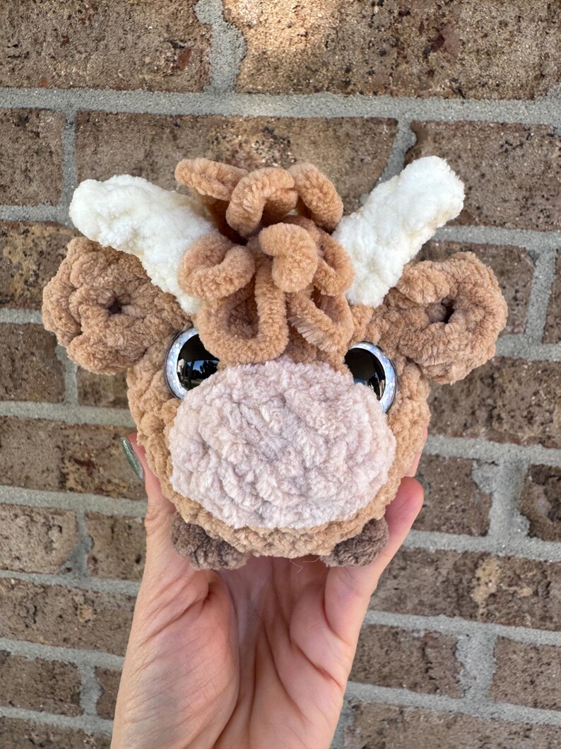 Puede incluir: Cabeza de vaca de peluche hecha a mano con ojos negros grandes y brillantes. La vaca tiene un cuerpo marr&oacute;n claro, una nariz color crema y cuernos color crema. El peluche se sostiene en una mano sobre un fondo de ladrillos.