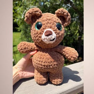 May include: A handmade brown teddy bear with large green eyes, a white muzzle, and a brown nose. The bear is crocheted with a textured yarn and has a smiling expression. The bear is approximately 8 inches tall.