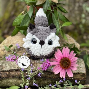 May include: A handmade crochet fox with gray, white, and black details. The fox is sitting on a log surrounded by flowers and greenery. A sticker with the text "The Cozy Buckeye" is visible.