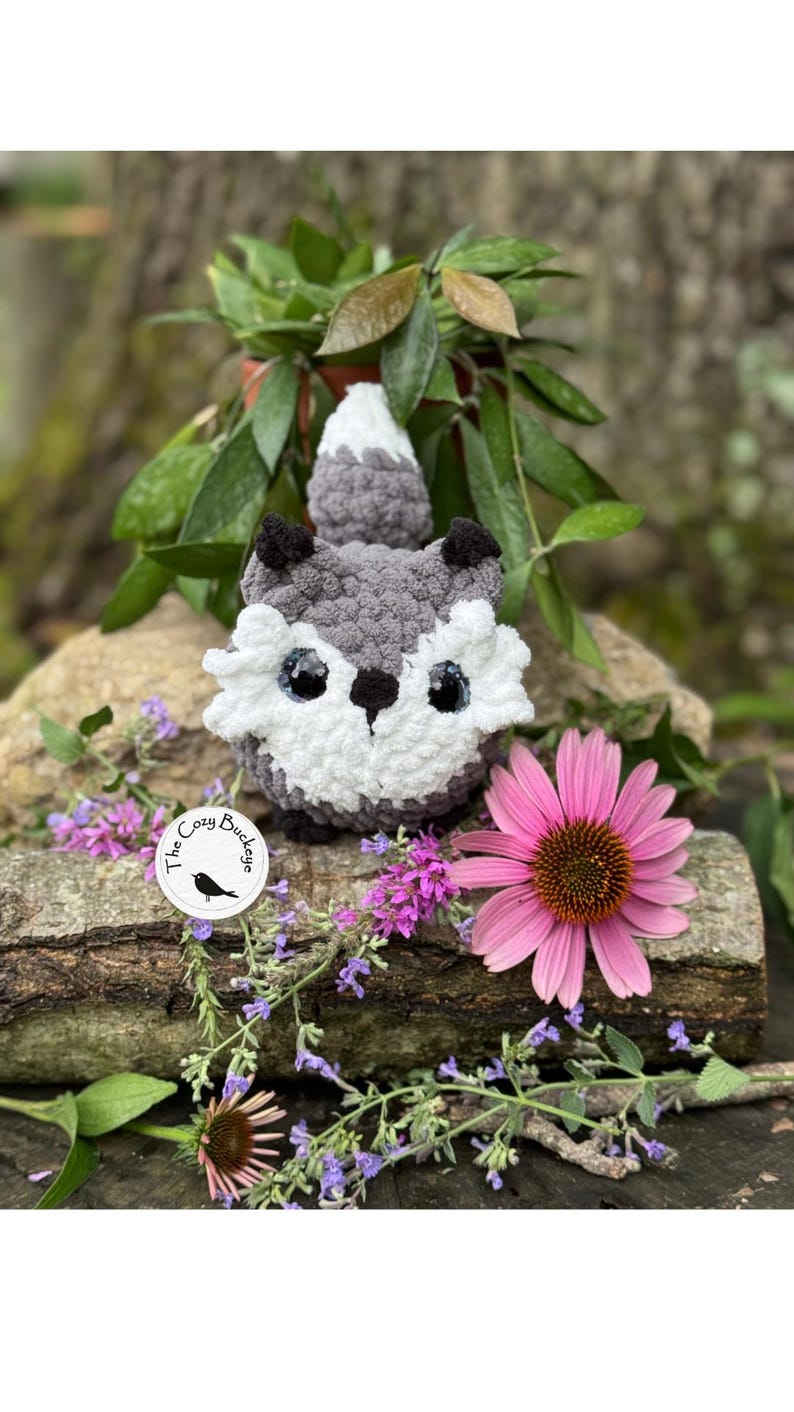 May include: A handmade crochet fox with gray, white, and black details. The fox is sitting on a log surrounded by flowers and greenery. A sticker with the text "The Cozy Buckeye" is visible.