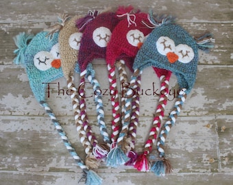 May include: Five crocheted owl hats with long braided tails in teal, brown, burgundy, purple, and blue. The hats have white eyes and orange beaks.
