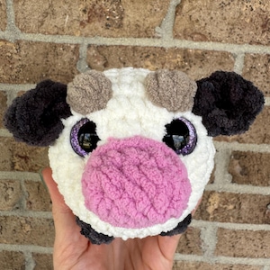 Puede incluir: Un juguete de vaca de peluche hecho a mano con cuerpo blanco, orejas negras y hocico rosa. La vaca tiene ojos grandes y brillantes de color p&uacute;rpura y peque&ntilde;os cuernos marrones. El juguete est&aacute; hecho de hilo suave y texturizado.