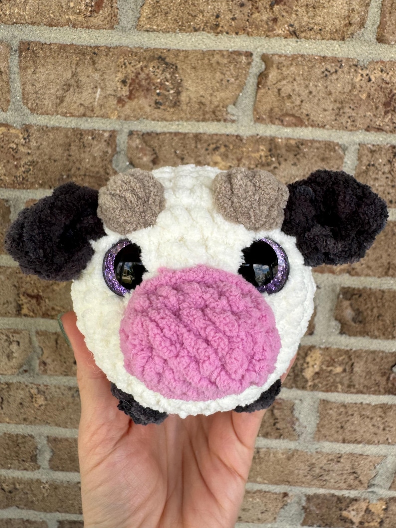 Puede incluir: Un juguete de vaca de peluche hecho a mano con cuerpo blanco, orejas negras y hocico rosa. La vaca tiene ojos grandes y brillantes de color p&uacute;rpura y peque&ntilde;os cuernos marrones. El juguete est&aacute; hecho de hilo suave y texturizado.