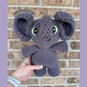 Puede incluir: Un elefante gris hecho a mano con ganchillo, con orejas grandes y ojos verdes brillantes. El elefante est&aacute; sostenido en la mano de una persona. El texto "@thecozybuckeye" es visible en la esquina superior izquierda. El fondo es una pared de ladrillos.
