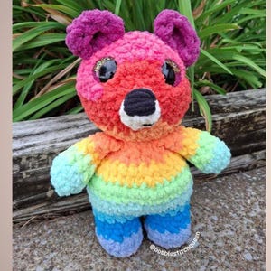 May include: A handmade, rainbow-colored crochet bear with a black nose, white mouth, and large, glittery eyes. The bear's fur transitions from pink to red, orange, yellow, green, and blue. The bear has purple ears and blue feet. The text "@bobblestitchqueen" is visible.