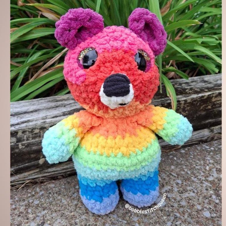 May include: A handmade, rainbow-colored crochet bear with a black nose, white mouth, and large, glittery eyes. The bear's fur transitions from pink to red, orange, yellow, green, and blue. The bear has purple ears and blue feet. The text "@bobblestitchqueen" is visible.