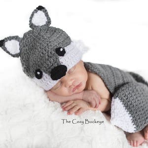 Pode incluir: Um recém-nascido vestindo um traje de lobo de crochê cinza e branco. O traje inclui um chapéu com orelhas de lobo e uma cauda. O bebê está dormindo em um cobertor branco e felpudo.