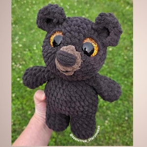 May include: A handmade black crochet bear with large, glittery gold eyes and a brown nose. The bear has a textured, plush appearance and is shown against a green background. The bear is a stuffed animal.