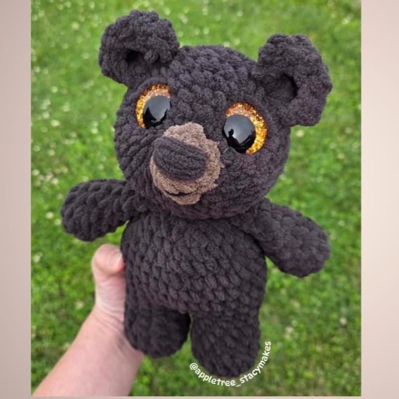 May include: A handmade black crochet bear with large, glittery gold eyes and a brown nose. The bear has a textured, plush appearance and is shown against a green background. The bear is a stuffed animal.