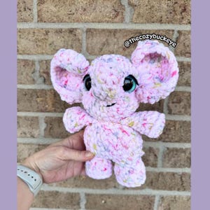 Puede incluir: Un juguete de elefante de peluche hecho a mano en tonos de rosa, blanco y amarillo. El elefante tiene orejas grandes y ca&iacute;das, ojos negros grandes con acentos turquesa y una peque&ntilde;a trompa. El texto "@thecozybuckeye" es visible. El juguete est&aacute; hecho de hilo suave y texturizado.