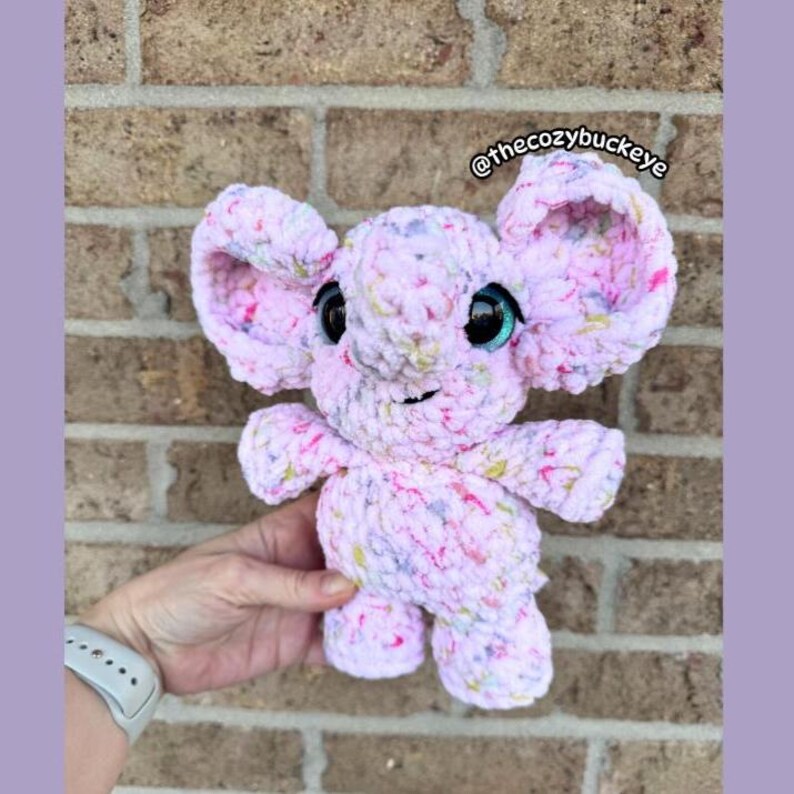 Puede incluir: Un juguete de elefante de peluche hecho a mano en tonos de rosa, blanco y amarillo. El elefante tiene orejas grandes y ca&iacute;das, ojos negros grandes con acentos turquesa y una peque&ntilde;a trompa. El texto "@thecozybuckeye" es visible. El juguete est&aacute; hecho de hilo suave y texturizado.