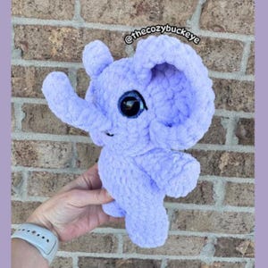 Puede incluir: Un elefante de ganchillo lavanda con ojos grandes, negros y azules. El elefante tiene una trompa, orejas y cuatro patas. El texto "@thecozybuckeye" es visible en la esquina superior derecha. El elefante es un art&iacute;culo hecho a mano.