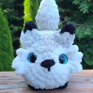 May include: A handmade plush toy in the shape of a white and gray creature with black accents. The toy has large blue eyes and a black nose. The plush is made of soft, textured yarn and sits on a wooden surface.