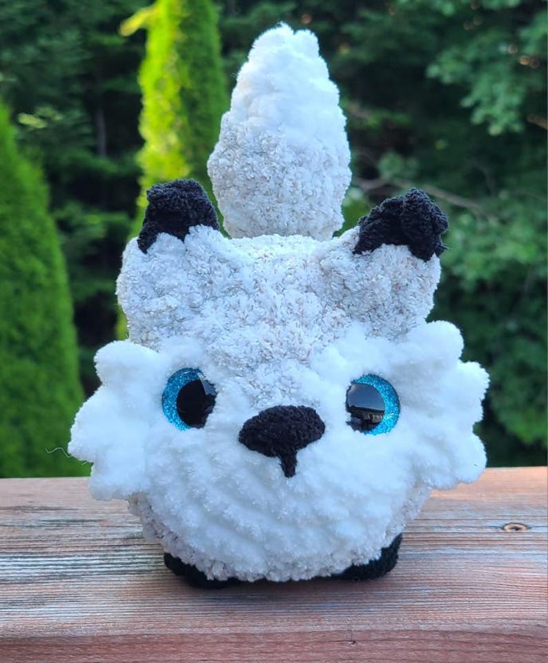 May include: A handmade plush toy in the shape of a white and gray creature with black accents. The toy has large blue eyes and a black nose. The plush is made of soft, textured yarn and sits on a wooden surface.
