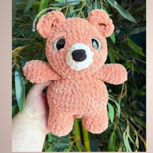 May include: A handmade, peach-colored teddy bear with large, expressive eyes and a black nose. The plush toy has a textured, fuzzy appearance and is designed with a classic bear shape, including rounded ears and limbs. The bear is a soft toy.