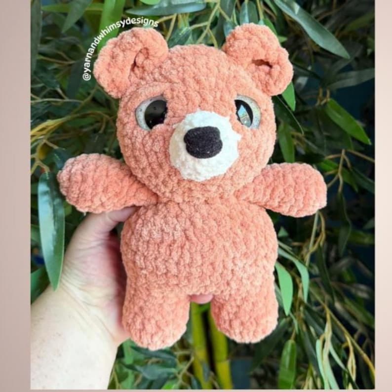May include: A handmade, peach-colored teddy bear with large, expressive eyes and a black nose. The plush toy has a textured, fuzzy appearance and is designed with a classic bear shape, including rounded ears and limbs. The bear is a soft toy.