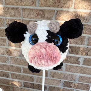 Puede incluir: Una cabeza de vaca de peluche en un palo, con un patr&oacute;n blanco y negro, ojos azules grandes y una nariz rosa. La vaca tiene orejas negras y peque&ntilde;os pompones marrones. El juguete se sostiene contra un fondo de ladrillos.