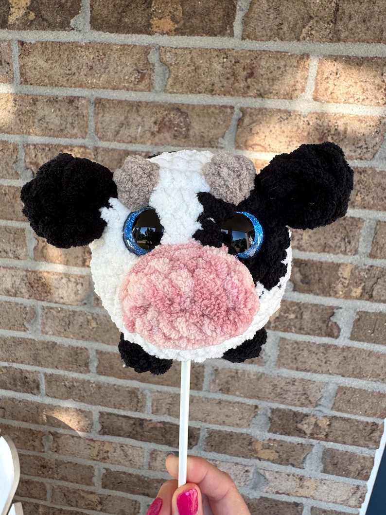 Puede incluir: Una cabeza de vaca de peluche en un palo, con un patr&oacute;n blanco y negro, ojos azules grandes y una nariz rosa. La vaca tiene orejas negras y peque&ntilde;os pompones marrones. El juguete se sostiene contra un fondo de ladrillos.