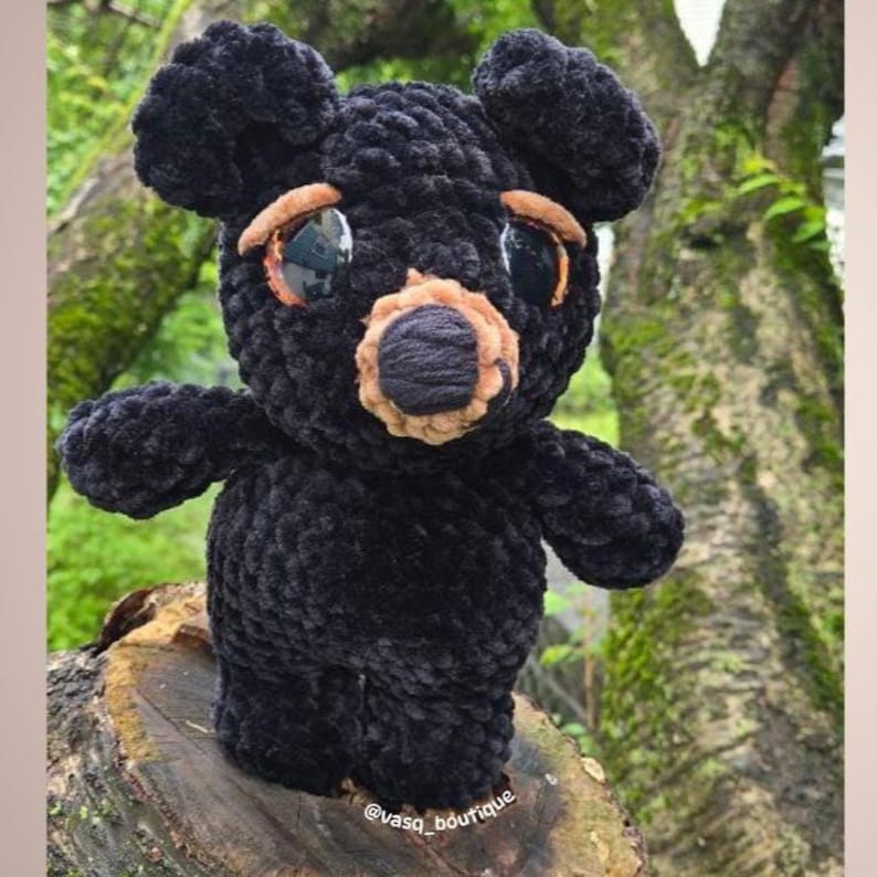 May include: A handmade black bear plush toy with large, expressive eyes and a tan snout. The bear is made of a soft, textured material and is standing on a wooden surface. The bear's eyes have a reflective quality.