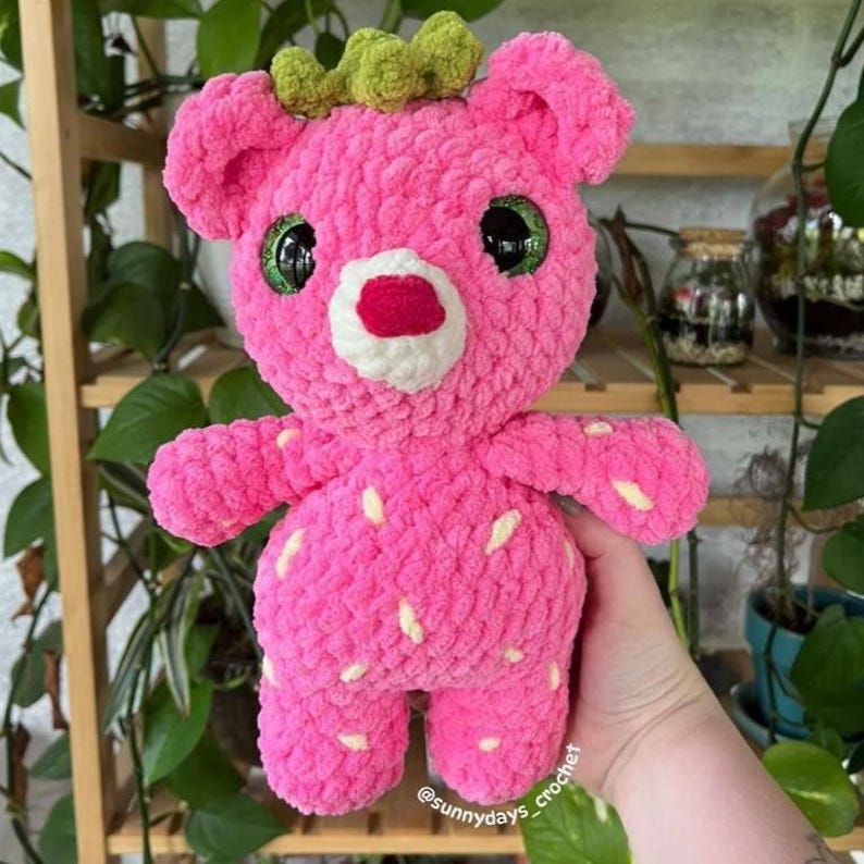 May include: A handmade pink crochet bear with a strawberry design. The plush toy has a white muzzle, red mouth, and green eyes. The bear's body is covered in white seed-like details, and it has a green crown. The text "@sunnydays_crochet" is visible.